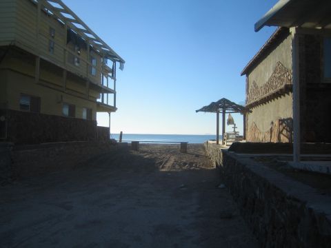 1990 Ford Bronco - sandy lot runs between two stone-trimmed buildings toward a calm blue sea under a clear sky.