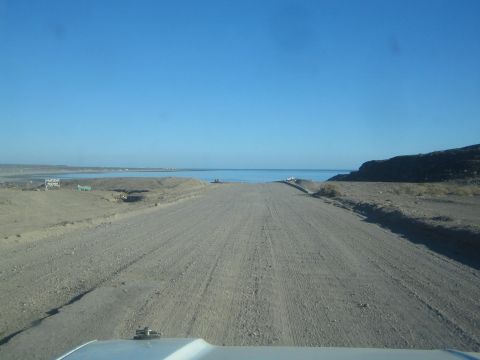1990 Ford Bronco - From behind a vehicle hood, a graded dirt road runs toward distant blue water and low hills under a clear sky.