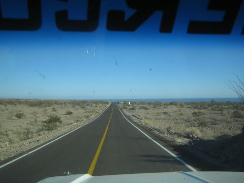 1990 Ford Bronco - Through a windshield, a paved two-lane highway with a yellow center line stretches across desert scrub toward a distant blue sea.