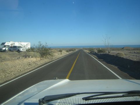 1990 Ford Bronco - Looking down a straight two-lane desert highway toward a blue sea, an RV and pickup parked at the roadside on the left.