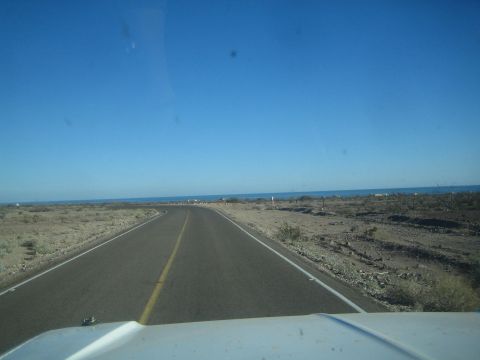 1990 Ford Bronco - driver's-eye view of an empty two-lane highway running through coastal desert scrub toward the blue ocean on the horizon.