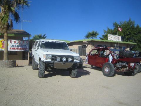 1990 Ford Bronco - white lifted truck with a roof light bar and front off-road lights tows a red tube-frame race truck on a dirt lot beside a SCORE emergency…