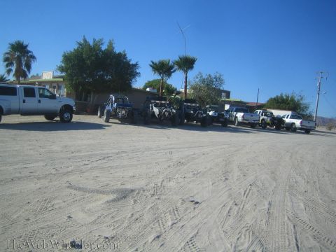 1990 Ford Bronco - Off-road buggies, trucks and a van line up on a sandy lot in front of low buildings and palm trees under a clear sky.