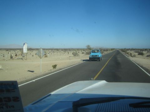 1990 Ford Bronco - small blue pickup drives toward the camera on a two-lane desert highway lined with a roadside cemetery and scrub.