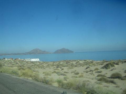 1990 Ford Bronco - Sandy dunes and a small white-building settlement front a calm blue bay backed by twin rounded mountain peaks.