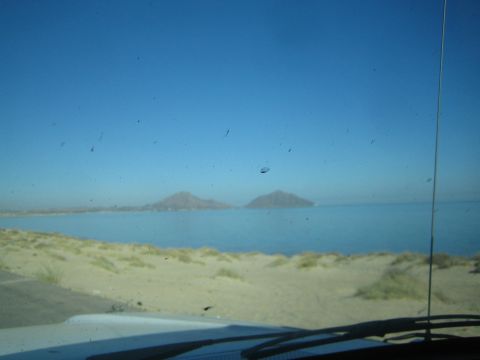 1990 Ford Bronco - Through a dusty windshield, a sandy beach meets a calm blue bay with two distant mountain peaks under clear sky.
