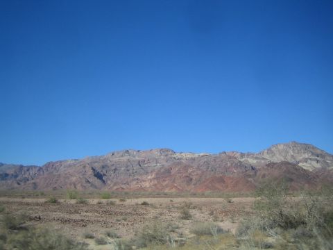 1990 Ford Bronco - long rugged ridge of reddish-brown desert mountains rises beyond sparse scrubland under a clear deep-blue sky.