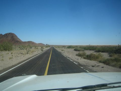 1990 Ford Bronco - patched two-lane highway stretches across flat open desert toward distant hills, seen over a light-colored hood.