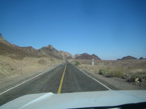 1990 Ford Bronco - two-lane highway curves through barren rocky desert hills under a clear blue sky, viewed over a vehicle hood.