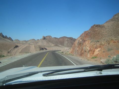 1990 Ford Bronco - two-lane road curves between jagged tan and rust-colored desert rock formations, viewed through a windshield with wiper.