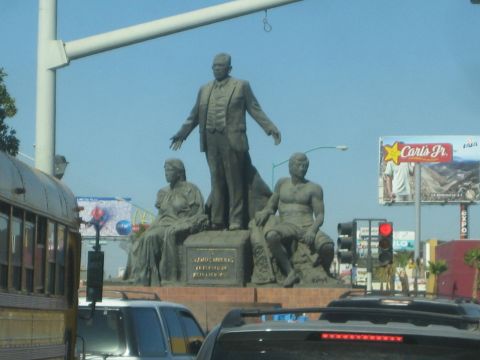 1990 Ford Bronco - bronze monument of a standing man flanked by two seated figures rises above busy street traffic near a Carl's Jr. billboard.