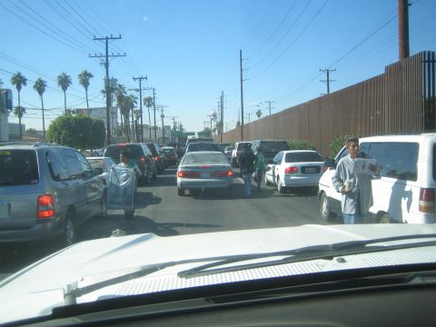 1990 Ford Bronco - Lanes of cars sit in heavy stop-and-go traffic beside a tall metal border wall as vendors walk between the vehicles.