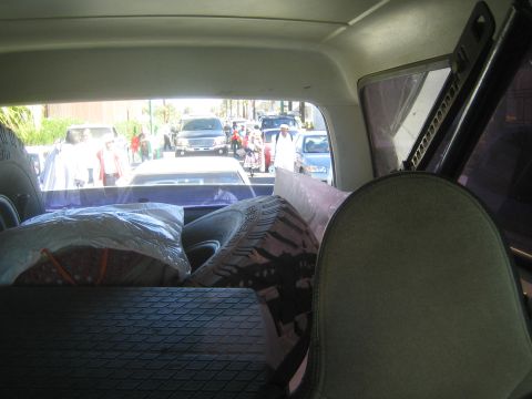 1990 Ford Bronco - From inside a vehicle's cargo area, blankets and gear sit ahead of an open rear hatch overlooking a crowded vendor-lined street.