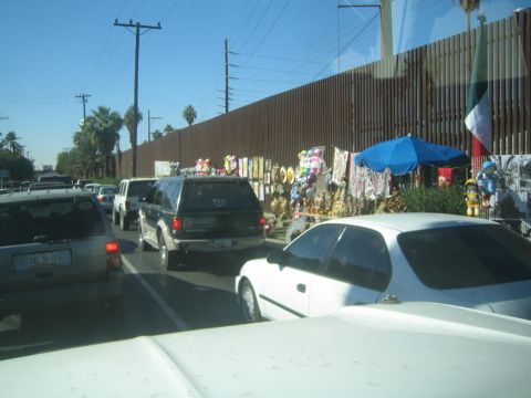 1990 Ford Bronco - Cars and an SUV queue in traffic beside a tall rust-colored border wall lined with souvenir stalls and a Mexican flag.