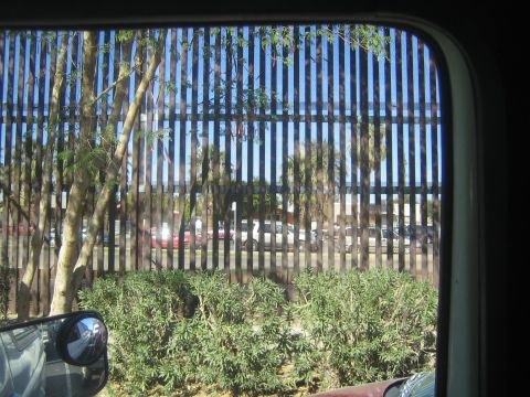 1990 Ford Bronco - Through a side window, a tall vertical steel-bar border fence separates the road from a parking lot, with the side mirror in frame.