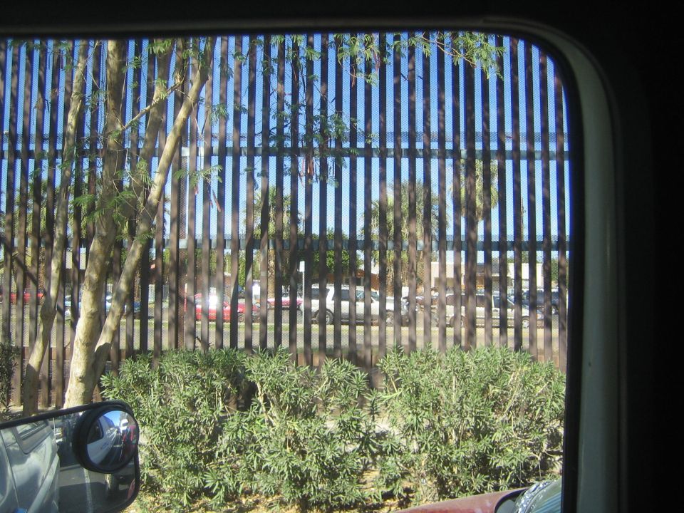 View through a vehicle window of a tall steel-bar border fence and a parking lot beyond