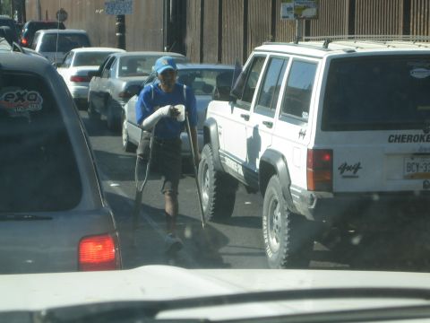 1990 Ford Bronco - man on crutches with one leg moves between stopped cars and a white Jeep Cherokee in border-area traffic beside the wall.