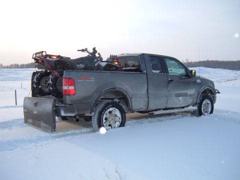 1979 Ford Bronco - A gray Ford F-150 pickup truck is stuck in the snow, with an ATV loaded in the bed and a plow attached to the rear.
