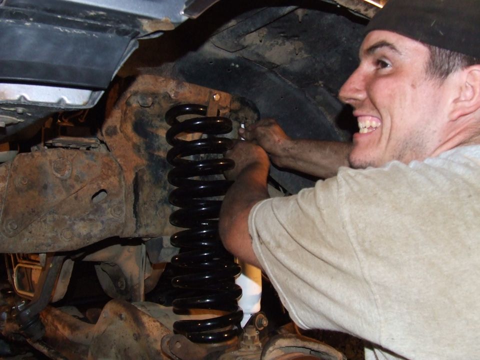 Man installing a black coil spring into a rusty vehicle suspension system