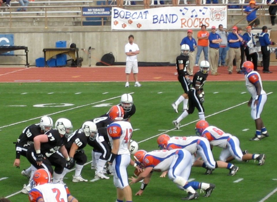 Football players in uniform on a field during a game, with spectators and a banner in the background