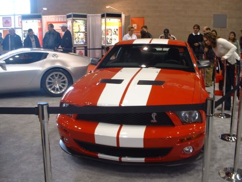 2000 Ford Crown Victoria - A red Ford Mustang Shelby GT500 with white racing stripes is displayed at an auto show, with a silver car and spectators in the background.
