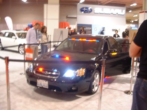 2000 Ford Crown Victoria - A black police car with its emergency lights on is displayed at an auto show, with a white car in the background.