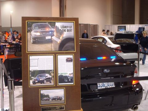 2000 Ford Crown Victoria - Subaru Legacy GT police car on display at an auto show, with informational board.