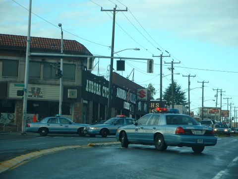 2000 Ford Crown Victoria - Police cruisers are seen on a street in front of businesses, including Aurora Cycle and Suzuki.