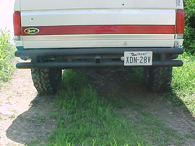 Rear of white Ford Bronco with red stripe, black tube bumper, Texas plate XDN-28V, on grassy dirt path