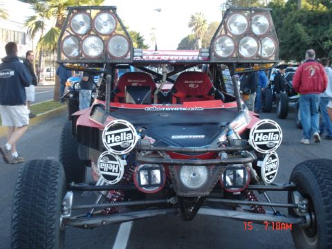 1990 Ford Bronco - red-and-black tube-frame off-road race buggy with two stacks of round roof lights and Hella lamps sits among spectators on a street.