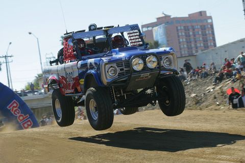 1990 Ford Bronco - blue-and-white early Ford Bronco-bodied off-road race truck catches air over a dirt course before a crowd, RedBull arch at left.