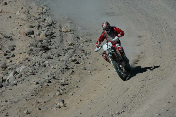 Dirt-bike racer in red gear riding fast through a gravelly desert wash