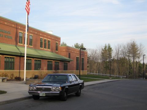 1991 Ford LTD Crown Victoria - A dark sedan is parked in front of a brick building with an American flag flying.