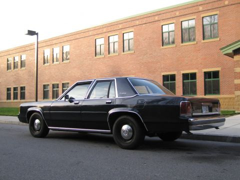 1991 Ford LTD Crown Victoria - A black sedan is parked on the street next to a curb, with a brick building in the background.