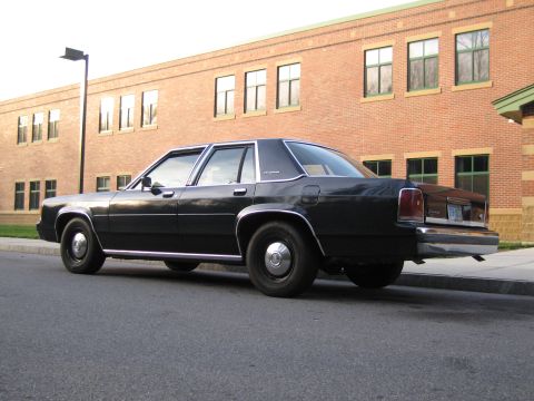 1991 Ford LTD Crown Victoria - This black sedan, likely from the 1980s, is parked curbside with a brick building behind it.