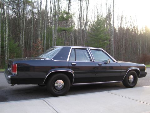 1991 Ford LTD Crown Victoria - Side view of a dark blue sedan with chrome trim, parked outdoors with a wooded area behind it.