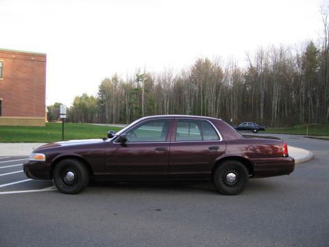 1991 Ford LTD Crown Victoria - A maroon sedan, likely a police vehicle, is parked on asphalt with a brick building and trees in the background.
