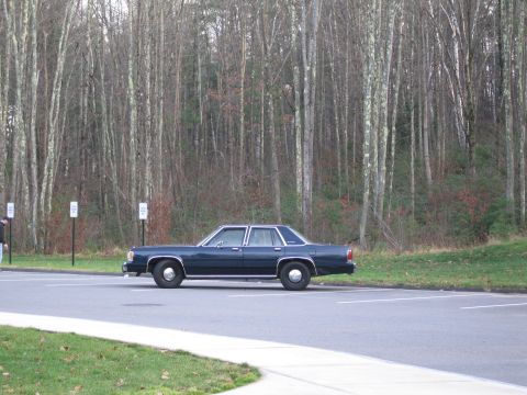 1991 Ford LTD Crown Victoria - A dark blue sedan is parked in a lot with a wooded area behind it.