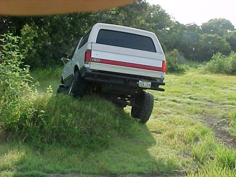 1990 Ford Bronco - White Ford Bronco with a red stripe on the tailgate is driving up a grassy incline.