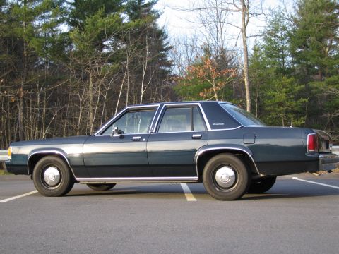 1991 Ford LTD Crown Victoria - A dark sedan with a "Crown Victoria" badge is seen from the side, parked outdoors.