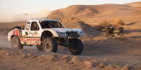 1990 Ford Bronco - white off-road race truck marked Collins Motorsports number 12 kicks up dust racing across desert terrain at dusk.