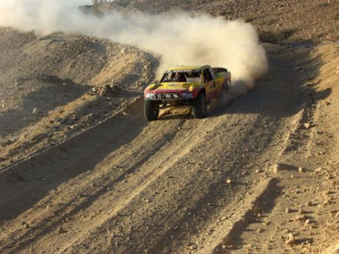 1990 Ford Bronco - yellow and red off-road race truck throws up a large dust cloud as it crests a dirt berm on a desert course.