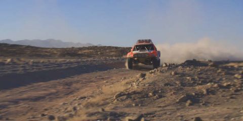 1990 Ford Bronco - red off-road race truck speeds across open desert terrain with hazy mountains on the horizon.
