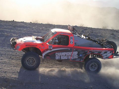 1990 Ford Bronco - red and white off-road race truck number 86 with Championship Off Road Racing graphics catches air on a rocky trail.