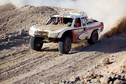 1990 Ford Bronco - white and red off-road race truck number 96 with Baldwin Motorsports graphics climbs a rocky desert hill.