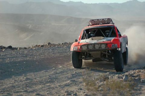 1990 Ford Bronco - red off-road race truck number 86 topped with a light bar drives down a desert trail toward the camera.