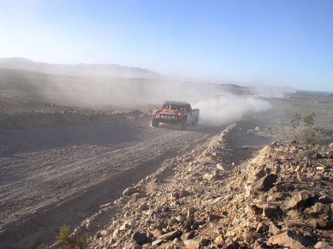 1990 Ford Bronco - red off-road race truck climbs a rocky desert grade, trailing a large cloud of dust under a clear sky.