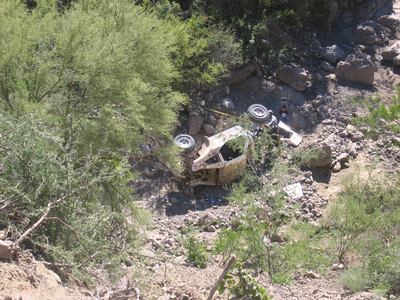 1990 Ford Bronco - white off-road vehicle lies upside down and wrecked among rocks and desert brush in a ravine.