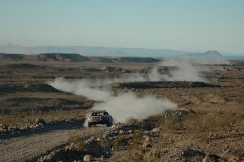 1990 Ford Bronco - off-road race truck kicks up a plume of dust as it rounds a curve on a wide desert course with distant hills.