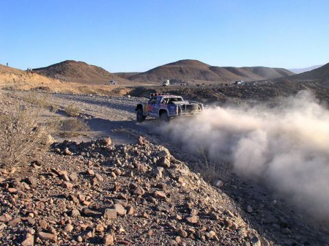 1990 Ford Bronco - off-road race truck disappears into a dust cloud on a rocky desert trail as spectators watch from the hills.